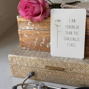 May include: A white card with black text and a simple line drawing of a rose. The text reads "I am stronger than the challenges I face". The card is sitting on a wooden box with a pink rose on top.