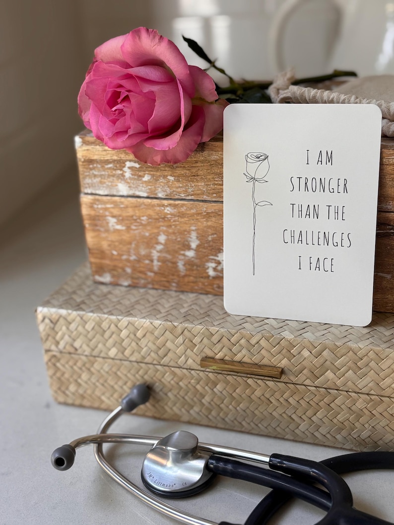May include: A white card with black text and a simple line drawing of a rose. The text reads "I am stronger than the challenges I face". The card is sitting on a wooden box with a pink rose on top.