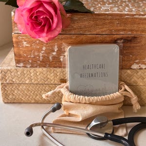 May include: A gray card with the text "Healthcare Affirmations" and a heart beat graphic. The card is in a small canvas bag with a stethoscope on a white surface.