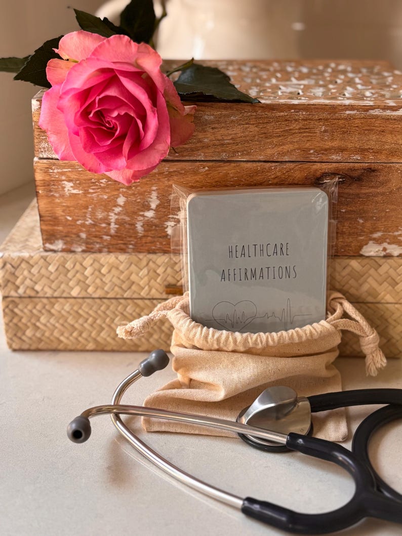 May include: A gray card with the text "Healthcare Affirmations" and a heart beat graphic. The card is in a small canvas bag with a stethoscope on a white surface.