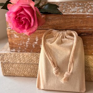 May include: A small, beige drawstring bag made of natural fabric with a knotted rope closure. The bag is sitting on top of a wooden box with a pink rose on top.