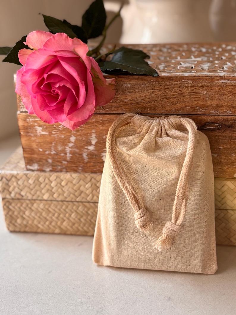 May include: A small, beige drawstring bag made of natural fabric with a knotted rope closure. The bag is sitting on top of a wooden box with a pink rose on top.