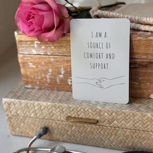 May include: A white card with the text "I am a source of comfort and support" and a simple line drawing of two hands holding each other. The card is sitting on a wooden box with a pink rose on top.