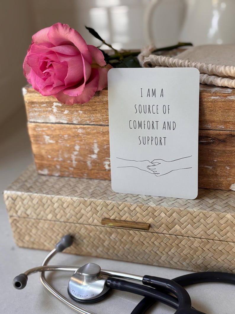 May include: A white card with the text "I am a source of comfort and support" and a simple line drawing of two hands holding each other. The card is sitting on a wooden box with a pink rose on top.