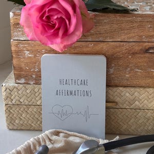 May include: A gray book with the text "Healthcare Affirmations" and a heart with a heartbeat line. The book is on top of a wooden box with a pink rose on top. A stethoscope is in the foreground.