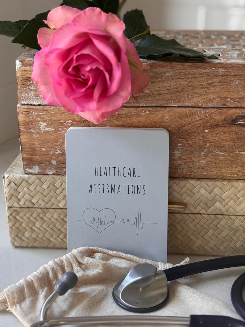 May include: A gray book with the text "Healthcare Affirmations" and a heart with a heartbeat line. The book is on top of a wooden box with a pink rose on top. A stethoscope is in the foreground.