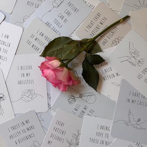 May include: A set of white affirmation cards with black line drawings and text. The cards feature positive messages for nurses and healthcare workers. The cards are scattered on a table with a pink rose in the center.