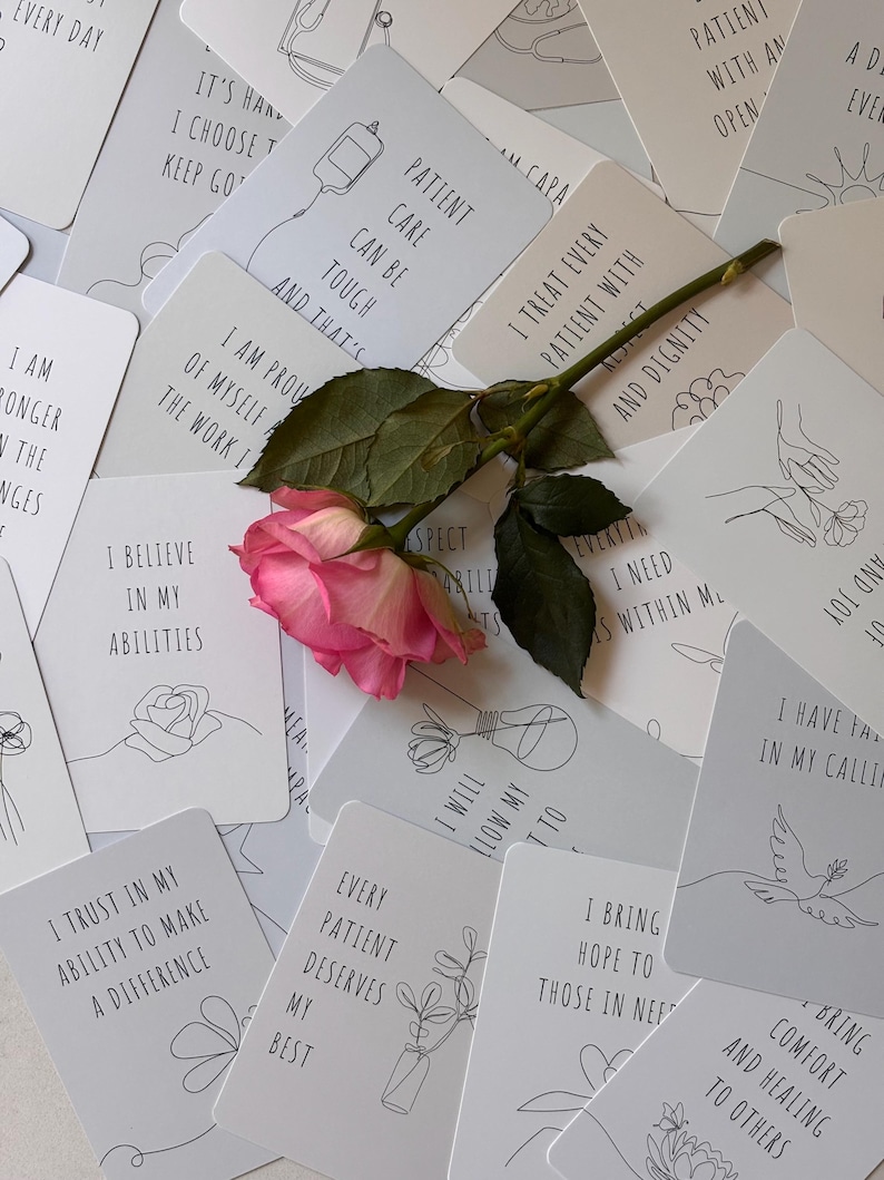 May include: A set of white affirmation cards with black line drawings and text. The cards feature positive messages for nurses and healthcare workers. The cards are scattered on a table with a pink rose in the center.