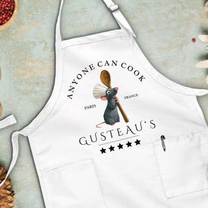 May include: White apron with a pocket and the text "ANYONE CAN COOK" in black. A cartoon rat holding a wooden spoon is in the center. The text "GUSTEAU'S" and five stars are below the rat.