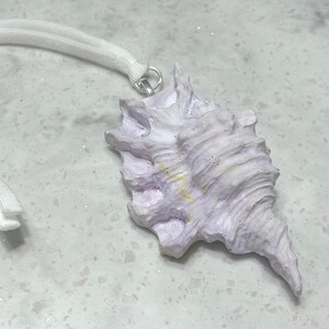 May include: A pale lavender seashell ornament featuring a textured, ridged surface with subtle yellow accents. The shell is attached to a silver-toned metal jump ring and a white elastic cord with a small white plastic fastener for hanging.