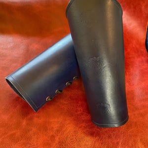 May include: A pair of dark blue leather bracers featuring a pointed top edge and embossed eagle motifs. The bracers are secured with black lacing threaded through metal eyelets along the side, providing a structured fit for the forearm.
