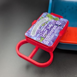 May include: A red plastic container with a label that reads "Concord Grape Jelly". The label is purple and white with a picture of grapes. The container is designed to hold a small amount of jelly.