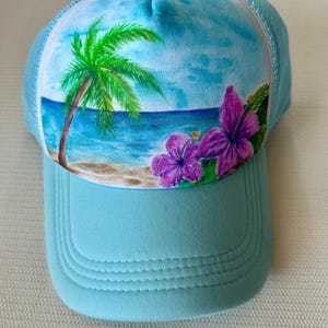 May include: A light blue trucker hat with a tropical beach scene. The hat features a painted design of a palm tree, ocean, and purple hibiscus flowers. The brim and front panel are solid light blue.