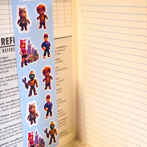 May include: A light blue bookmark featuring the Roblox logo at the top and a repeating vertical pattern of various cartoon characters, including a robot with a wrench, a figure in a red cap, and a castle, placed inside an open lined notebook.