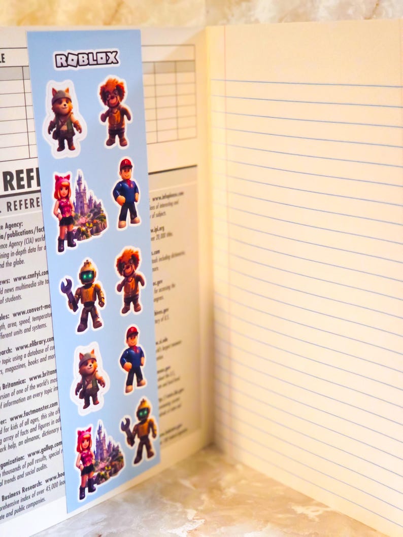 May include: A light blue bookmark featuring the Roblox logo at the top and a repeating vertical pattern of various cartoon characters, including a robot with a wrench, a figure in a red cap, and a castle, placed inside an open lined notebook.