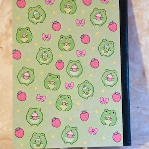 May include: A notebook with a light green cover featuring a repeating pattern of small green frogs, pink strawberries, pink bows, and yellow stars. The frogs are depicted holding either a pink cupcake, a heart, or a boba tea drink. The spine is black.