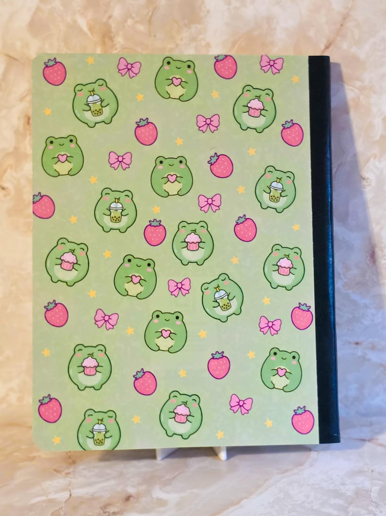 May include: A notebook with a light green cover featuring a repeating pattern of small green frogs, pink strawberries, pink bows, and yellow stars. The frogs are depicted holding either a pink cupcake, a heart, or a boba tea drink. The spine is black.