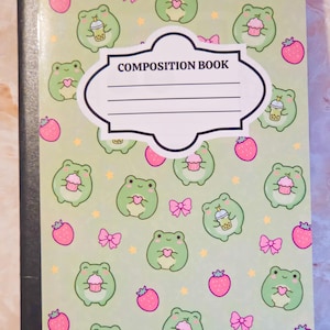May include: A light green composition book featuring a repeating pattern of small green frogs, pink strawberries, and pink bows. The frogs are depicted holding either a pink cupcake or a boba tea drink. A white label with the text COMPOSITION BOOK is centred.