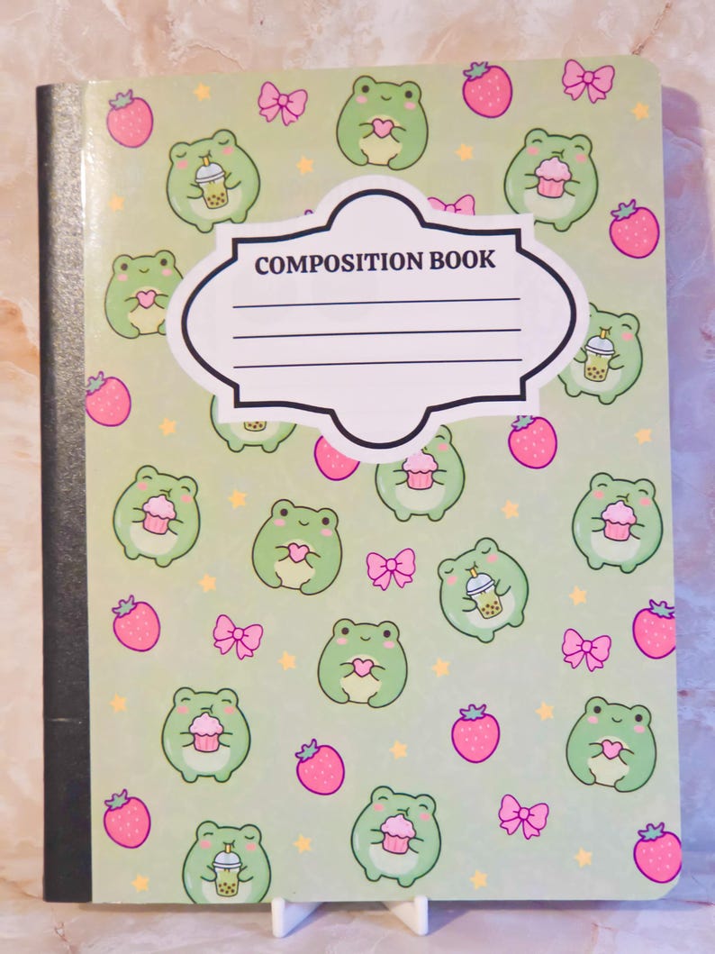 May include: A light green composition book featuring a repeating pattern of small green frogs, pink strawberries, and pink bows. The frogs are depicted holding either a pink cupcake or a boba tea drink. A white label with the text COMPOSITION BOOK is centred.