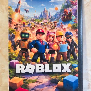 May include: A rectangular notebook featuring a colourful Roblox graphic on the front cover. The image displays five blocky characters standing in a grassy landscape with a castle in the background, a rocket, and the white Roblox logo at the bottom.