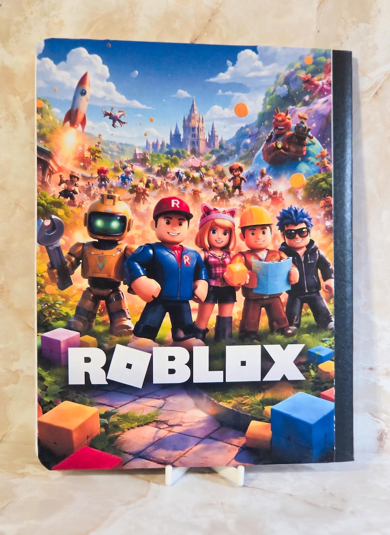 May include: A rectangular notebook featuring a colourful Roblox graphic on the front cover. The image displays five blocky characters standing in a grassy landscape with a castle in the background, a rocket, and the white Roblox logo at the bottom.