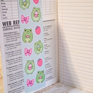 May include: A light blue bookmark featuring a repeating pattern of green frogs, pink strawberries, pink bows, and frogs holding bubble tea or cupcakes. The bookmark is placed inside an open notebook with lined pages and a printed class schedule.
