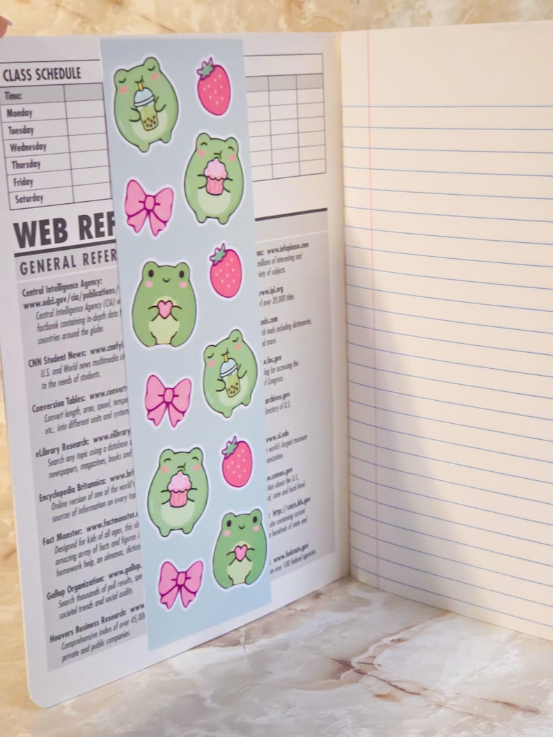 May include: A light blue bookmark featuring a repeating pattern of green frogs, pink strawberries, pink bows, and frogs holding bubble tea or cupcakes. The bookmark is placed inside an open notebook with lined pages and a printed class schedule.