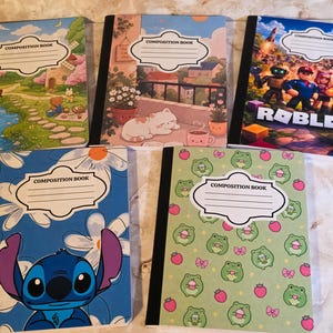 Peut inclure: Cinq cahiers de composition avec des reliures noires et des étiquettes blanches portant la mention COMPOSITION BOOK. Les motifs incluent un paysage avec un pont, un chat sur un balcon, une scène Roblox, un personnage bleu et des motifs de grenouilles.
