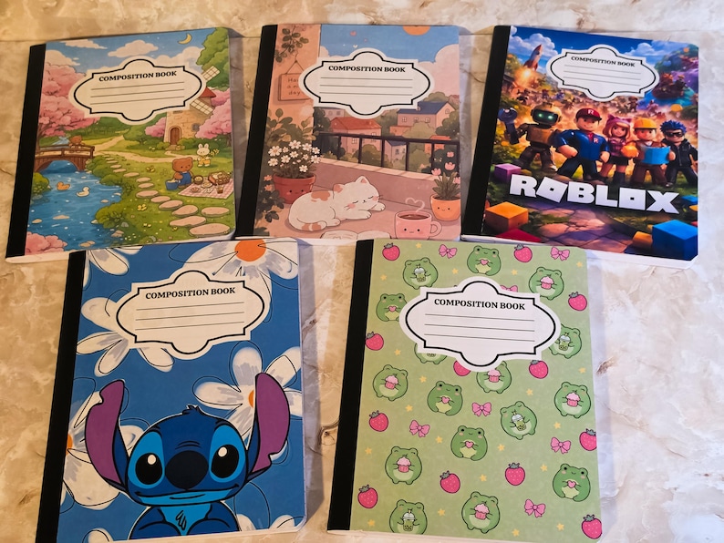 May include: Five composition books with black spines and white labels reading COMPOSITION BOOK. Designs include a landscape with a bridge, a cat on a balcony, a Roblox scene, a blue character with flowers, and a green pattern with frogs and strawberries.