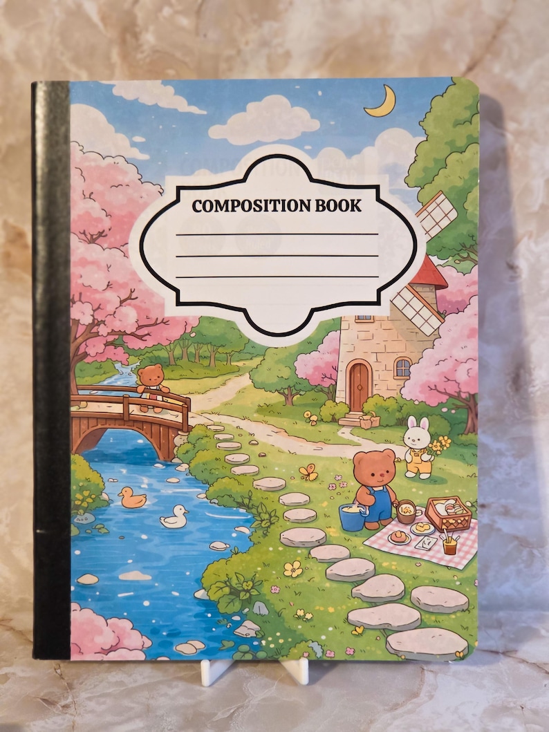 May include: A composition book featuring a colourful illustration of a landscape with a stream, a wooden bridge, a stone windmill, and cartoon bears and a rabbit having a picnic. The cover includes a white label with the text COMPOSITION BOOK.