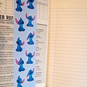 May include: A vertical light blue bookmark featuring a repeating pattern of the blue alien character Stitch with large ears. The bookmark is placed inside an open notebook with lined, cream-coloured pages and a page containing a printed web reference list.