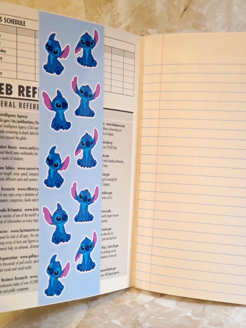 May include: A vertical light blue bookmark featuring a repeating pattern of the blue alien character Stitch with large ears. The bookmark is placed inside an open notebook with lined, cream-coloured pages and a page containing a printed web reference list.