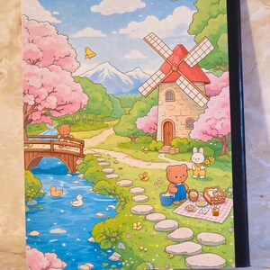 May include: A notebook with a colourful illustration featuring a stone windmill, a blue river with ducks, a wooden bridge, and cherry blossom trees. A brown bear and a white rabbit are depicted in a green landscape with a stone path and picnic.