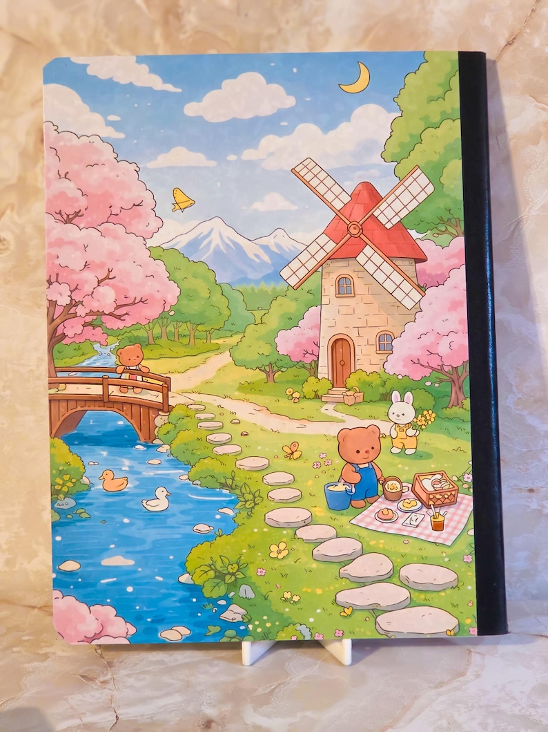 May include: A notebook with a colourful illustration featuring a stone windmill, a blue river with ducks, a wooden bridge, and cherry blossom trees. A brown bear and a white rabbit are depicted in a green landscape with a stone path and picnic.