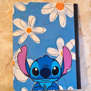 May include: A rectangular notebook featuring a blue cover with a repeating pattern of white daisies with orange centres. The character Stitch, a blue creature with large pink ears and black eyes, is positioned at the bottom of the cover.