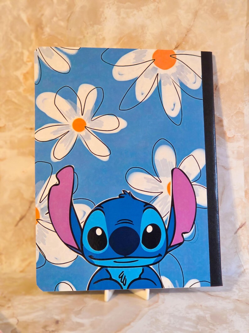 May include: A rectangular notebook featuring a blue cover with a repeating pattern of white daisies with orange centres. The character Stitch, a blue creature with large pink ears and black eyes, is positioned at the bottom of the cover.