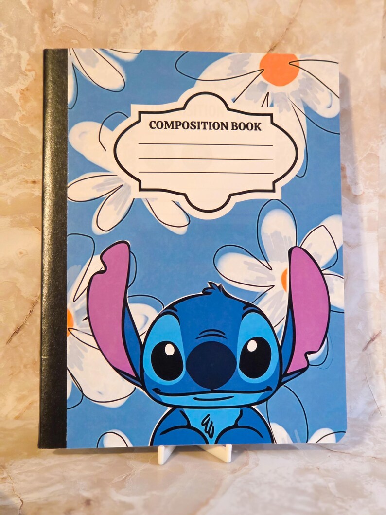 May include: A composition book with a blue cover featuring a repeating white daisy pattern with orange centres. The character Stitch is printed at the bottom with large purple ears. A white label with the text COMPOSITION BOOK is centred.