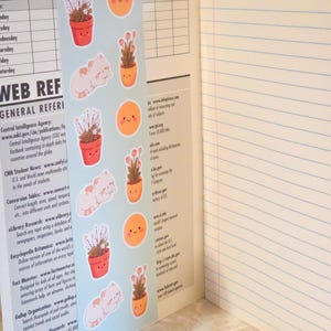 May include: A light blue bookmark featuring repeating illustrations of potted plants, sleeping white cats, and smiling orange suns, placed inside an open notebook with lined pages and a printed class schedule and web reference guide on the left side.