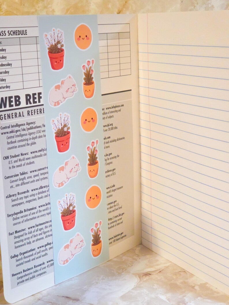 May include: A light blue bookmark featuring repeating illustrations of potted plants, sleeping white cats, and smiling orange suns, placed inside an open notebook with lined pages and a printed class schedule and web reference guide on the left side.
