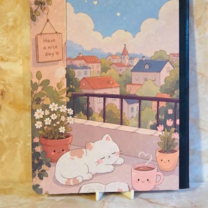 May include: A rectangular notebook featuring a colourful illustration of a white cat sleeping on a balcony. The scene includes potted plants, a steaming pink mug, and a sign that reads Have a nice day. The background shows a town with houses.