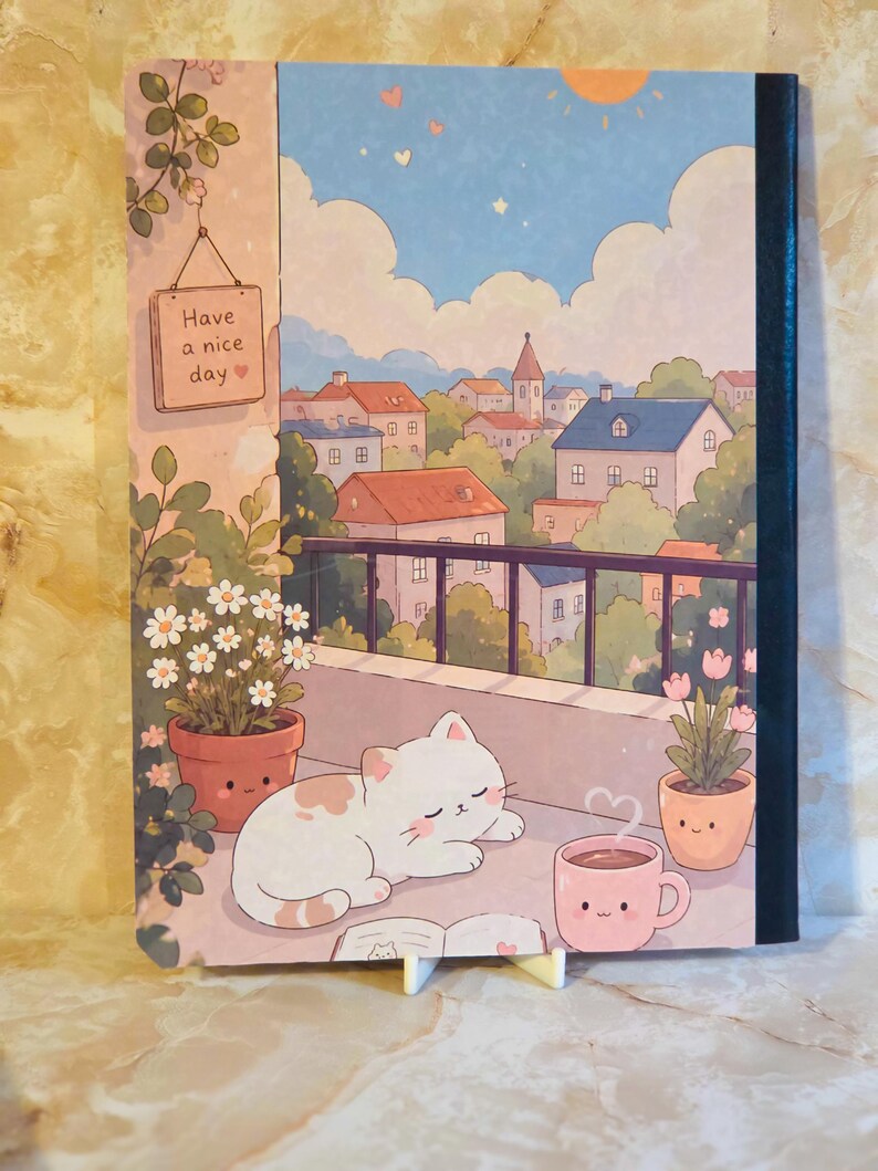 May include: A rectangular notebook featuring a colourful illustration of a white cat sleeping on a balcony. The scene includes potted plants, a steaming pink mug, and a sign that reads Have a nice day. The background shows a town with houses.