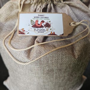 May include: A tan burlap drawstring bag with a rectangular cardboard tag attached by a string. The tag features a cartoon worm and rabbit, with text reading: RABBIT WORM CASTINGS, MADE FROM RABBIT DROPPINGS, 100% ORGANIC SOIL ENRICHMENT, NPK BOOST.