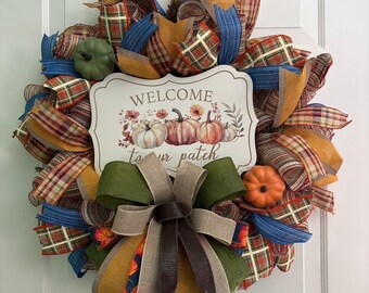 May include: A fall-themed wreath with a sign that reads "Welcome to your patch." The wreath features a variety of ribbons in plaid, blue, orange, and brown. Decorative pumpkins and a large bow complete the design. Perfect for autumn decor.