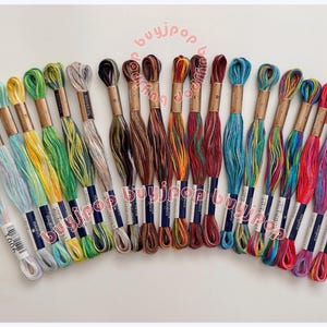 May include: A collection of colorful embroidery floss skeins arranged in a semi-circle. The floss is in various shades of blue, green, yellow, brown, red, and purple. Each skein is wrapped with a paper band and labeled. The background is white.