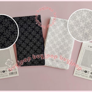 Può includere: Due pezzi di tessuto piegati, uno nero e uno bianco, con un motivo a croce ripetuto. Sono presenti anche due carte più piccole con un motivo simile. Il tessuto e le carte sono su una superficie rosa.