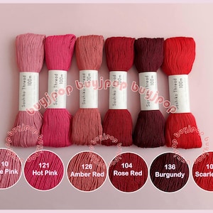 Könnte beinhalten: Eine Sammlung von sechs Spulen Sashiko-Garn in verschiedenen Rot- und Rosatönen. Jede Spule ist mit der Garnfarbe und -länge beschriftet. Die Farben reichen von Rose Pink bis Scharlachrot.