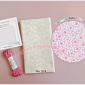 Può includere: Un kit per ricamo sashiko composto da un pannello di tessuto bianco con un motivo a fiori di ciliegio grigio chiaro, una matassina di filo da ricamo rosa variegato e una scheda di istruzioni. Un campione circolare mostra il ricamo finito.