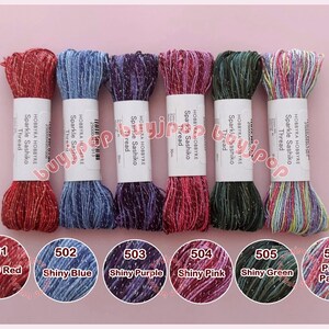 May include: Six spools of Hobbyra Hobbyre Sparkle Sashiko thread in various colors, including shiny red, blue, purple, pink, green, and a prism palette. Each spool is labeled with its color and number. The threads are displayed against a pink background.