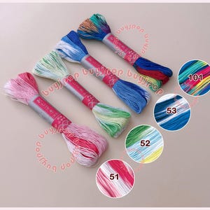 May include: Four skeins of variegated embroidery floss arranged diagonally. Each skein features a gradient of colors including pink and white, green and white, blue and white, and a multi-colored blend. Each has a paper label with Japanese text.