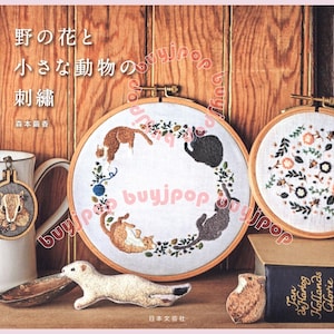 Puede incluir: Una exhibición que presenta aros de bordado con diseños de gatos y flores, un libro con texto japonés y un colgante de ardilla. Los aros de bordado se colocan contra un fondo de madera. También hay un pequeño animal de peluche.